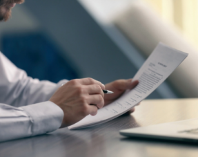Image of a man sat at a table reading a document with a pen in his hand and a laptop nearby.