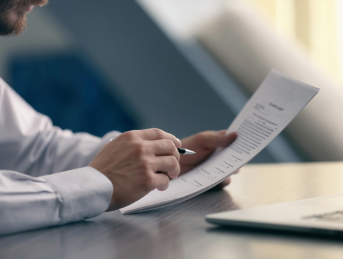 Image of a man sat at a table reading a document with a pen in his hand and a laptop nearby.