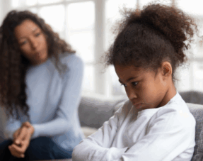 Image of a young child with her arms crossed looking upset with her mother sat nearby.