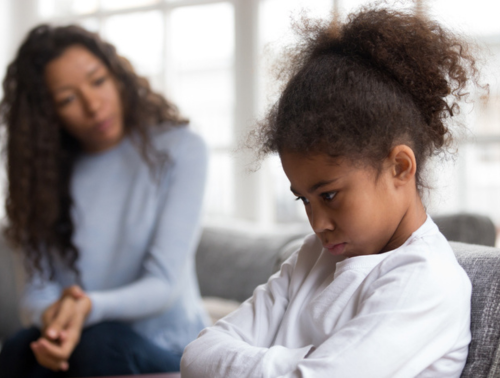 Image of a young child with her arms crossed looking upset with her mother sat nearby.