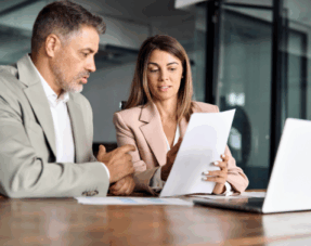 Image of a woman showing a document to a man with a laptop in front of them on a table.