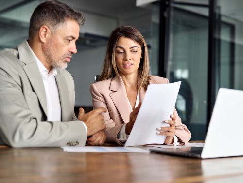 Image of a woman showing a document to a man with a laptop in front of them on a table.